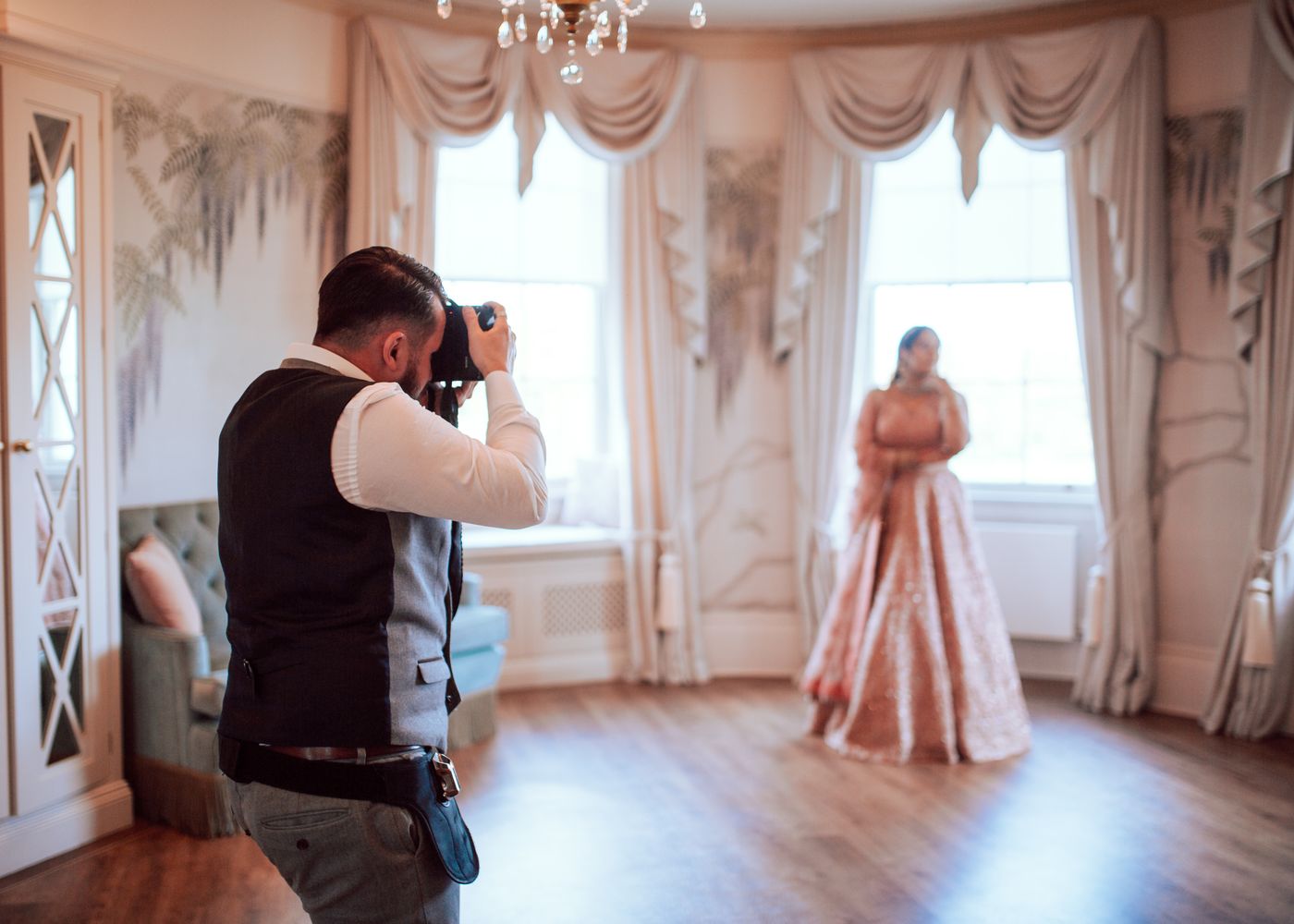 Ein Fotograf in Weste fotografiert eine Braut in einem rosa bestickten Kleid an einem hohen Fenster