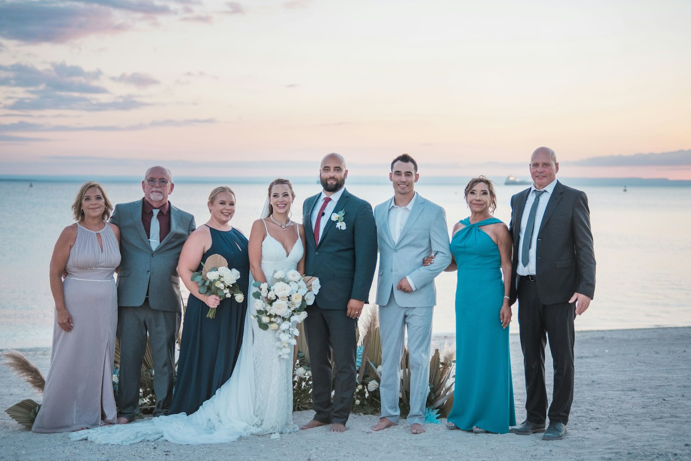 Una pareja de novios en la playa al atardecer con seis familiares vestidos de fiesta, la novia con un ramo blanco