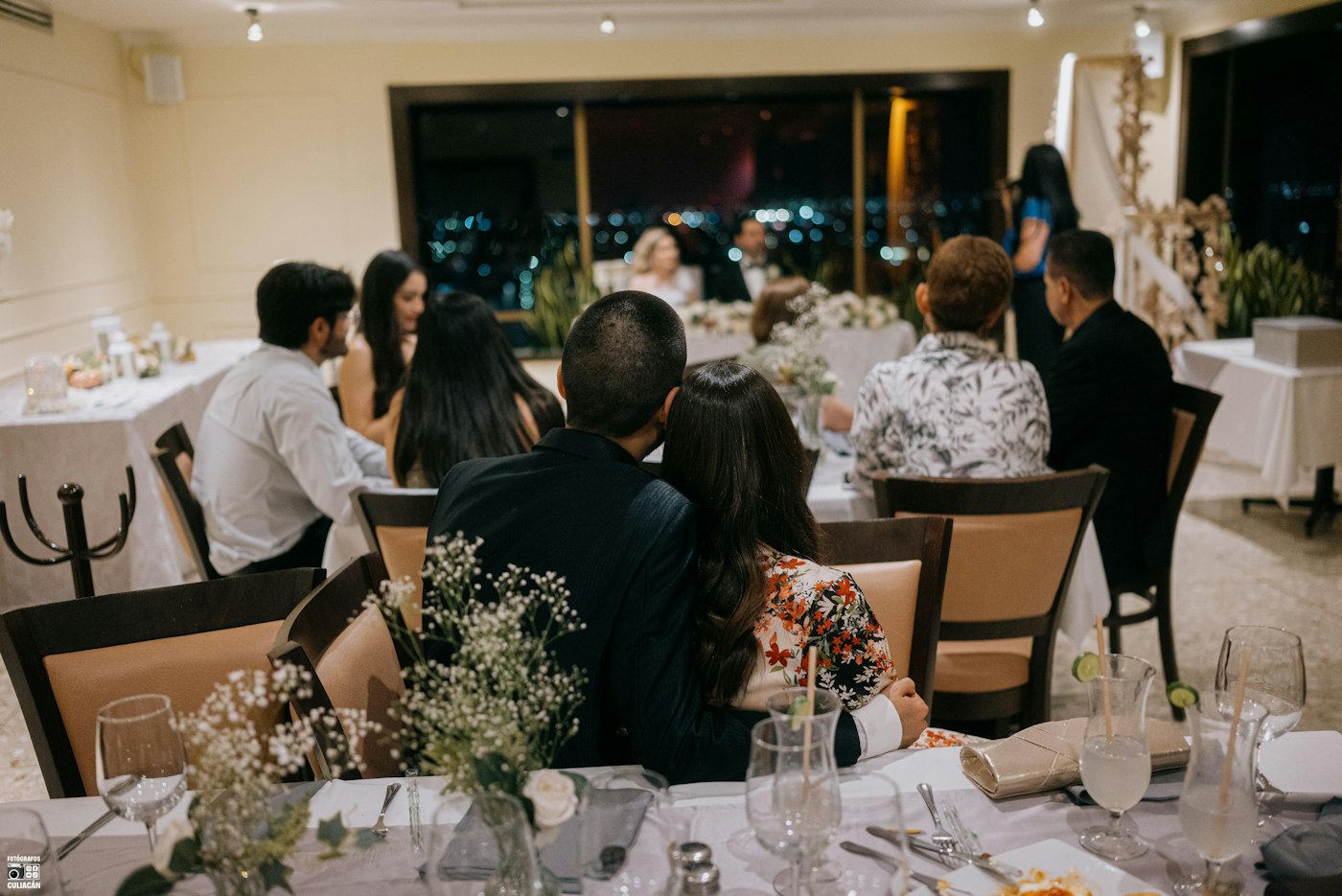 Una pequeña celebración de boda por la noche en un restaurante, los invitados en unas pocas mesas redondas con paniculata en jarrones de cristal y los novios en su mesa junto a la ventana