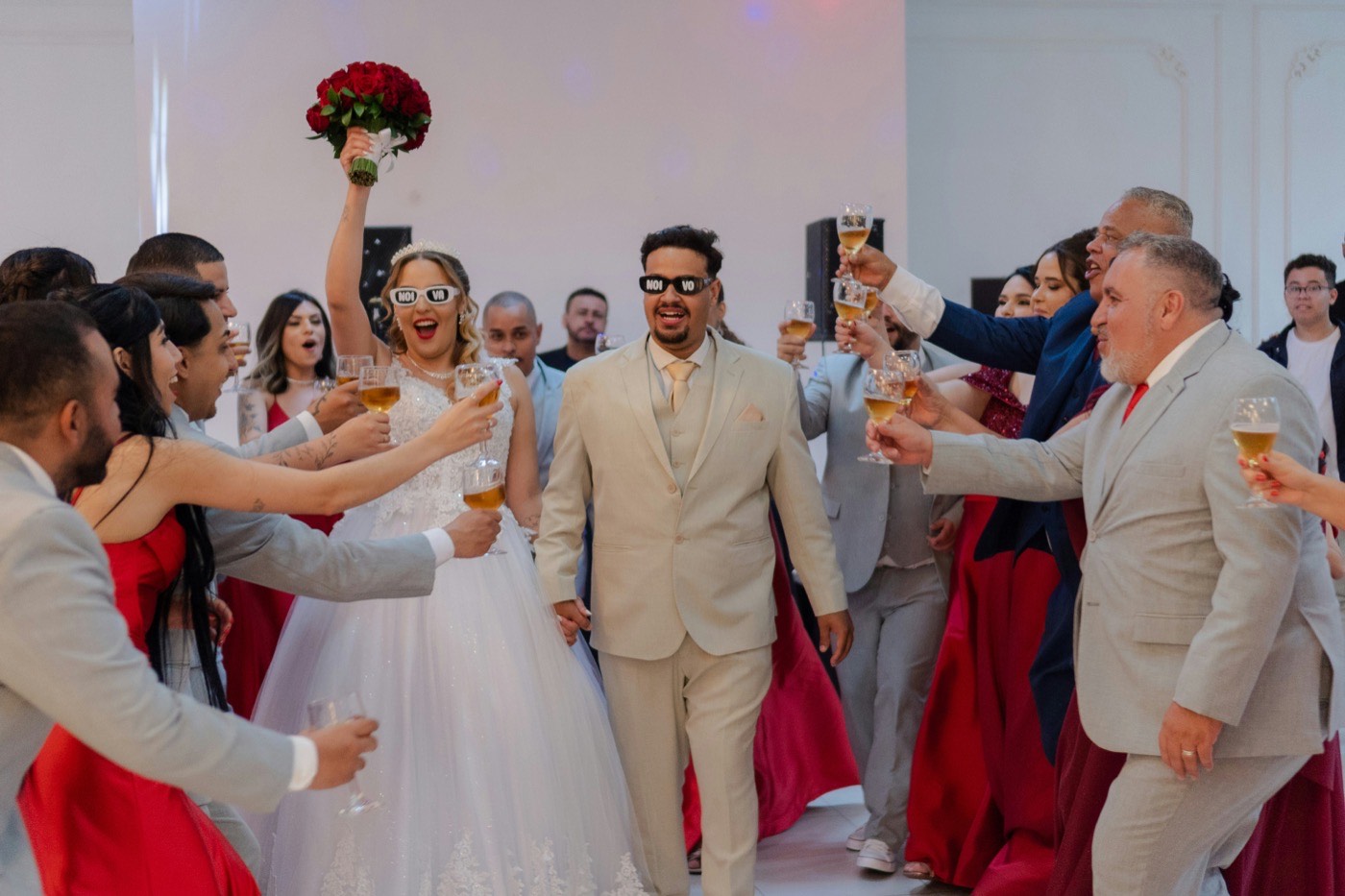 Guests raise glasses of champagne around the bride and groom as they walk in, the bride holding red roses above her head
