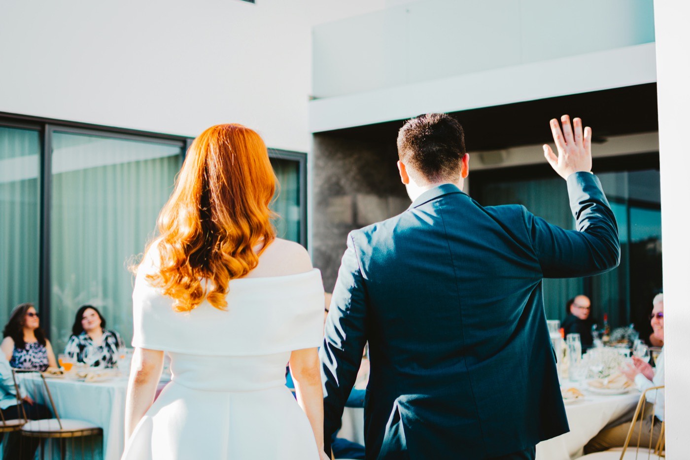 The bride and groom walk hand in hand towards a small group of guests at round tables on a sunny terrace