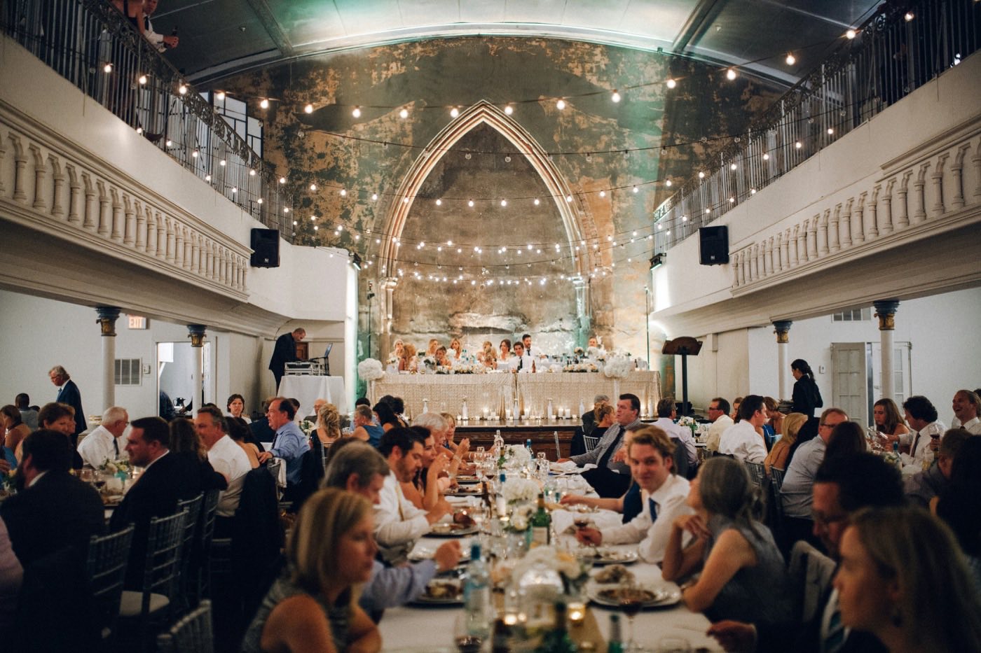 Guests eat dinner at long tables under string lights in an old church hall, with the wedding party at the head table