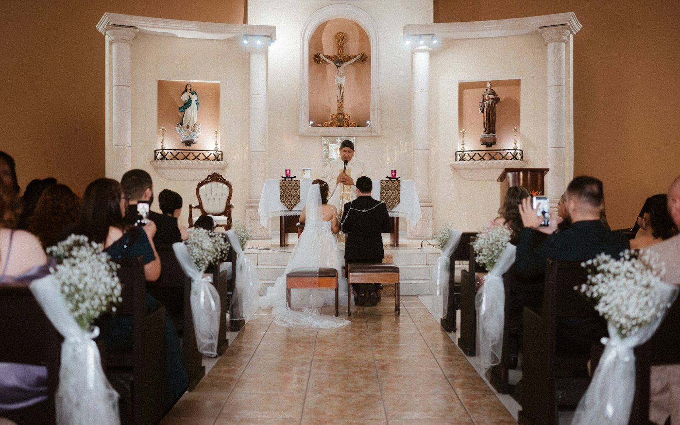 Guests in wooden pews decorated with white tulle bows and bunches of baby's breath, with the couple kneeling at the altar