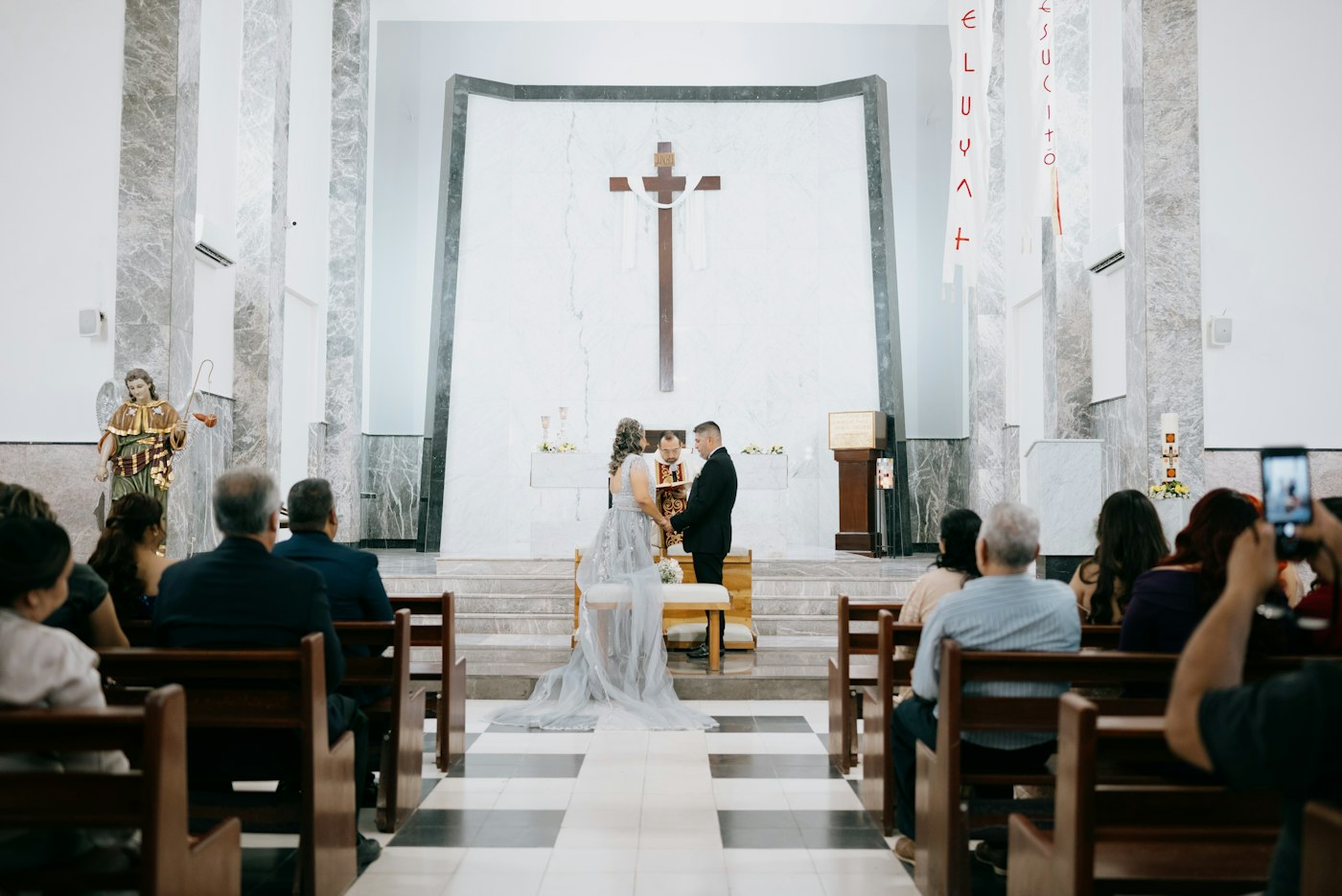 A couple holding hands at the altar of a white church with marble walls, guests watching from the pews