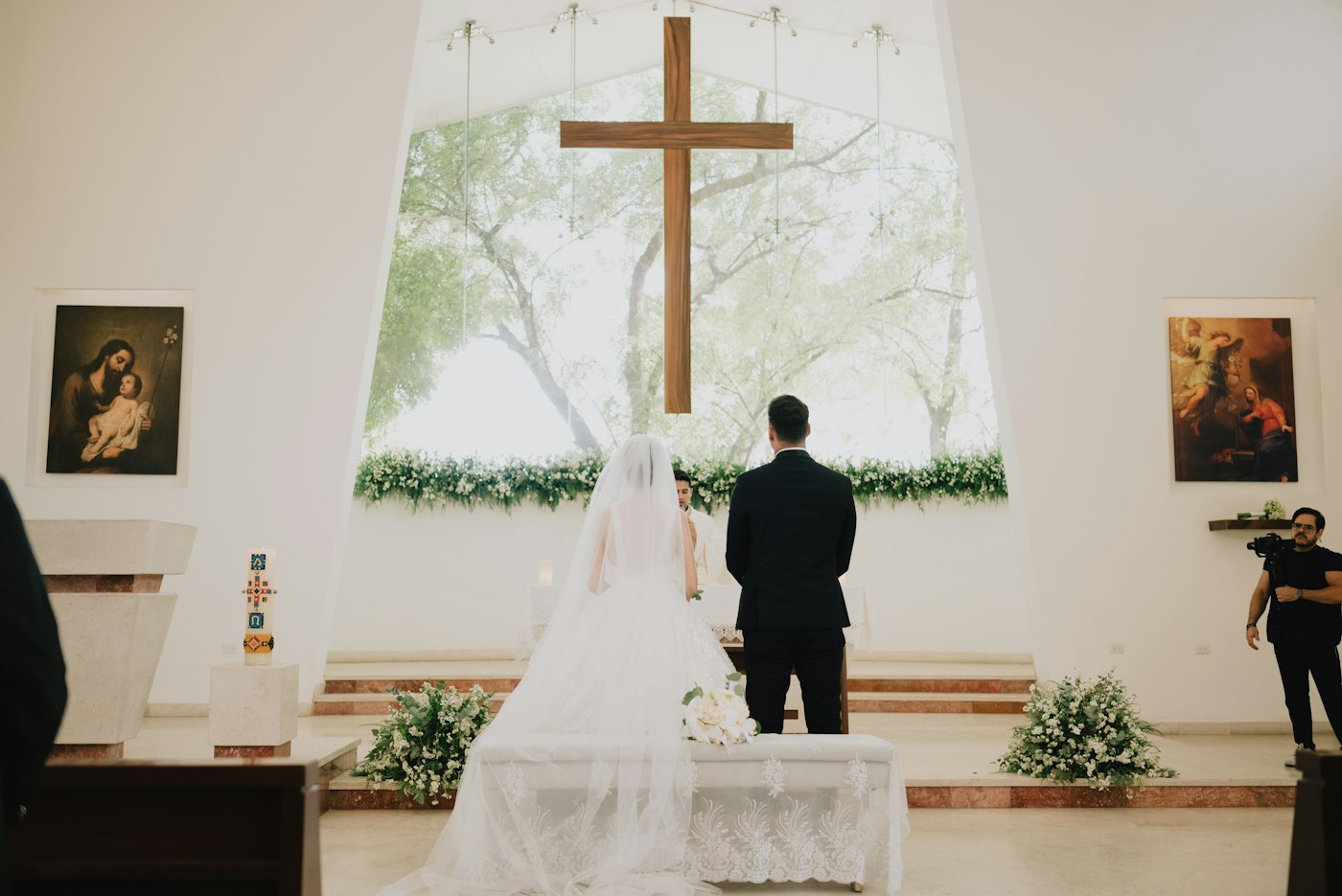 A bride and groom stand at a bright church altar with a garland of white flowers and greenery below a tall window and two flower arrangements on the steps