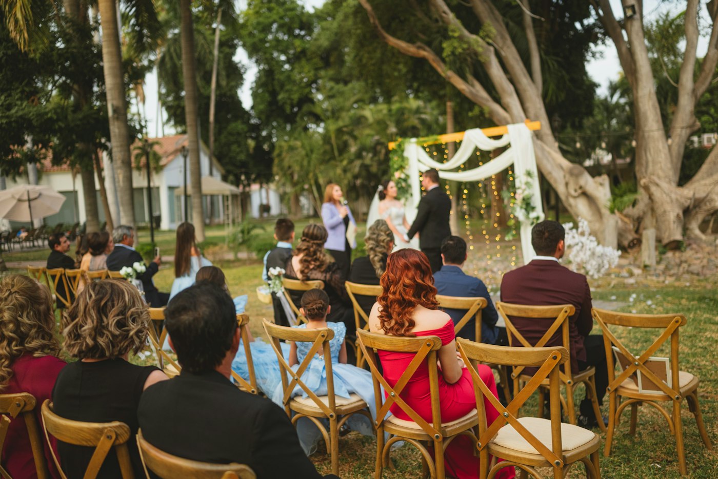 Guests on wooden chairs watch a small garden wedding under a draped fabric arch hung with fairy lights