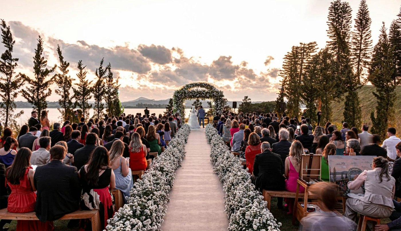 Guests on wooden benches watch an outdoor wedding ceremony at sunset, with an aisle lined in white flowers leading to a floral arch by a lake