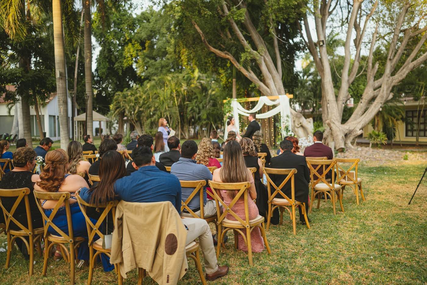 Guests on wooden folding chairs watching a couple stand under a white draped arch in a garden