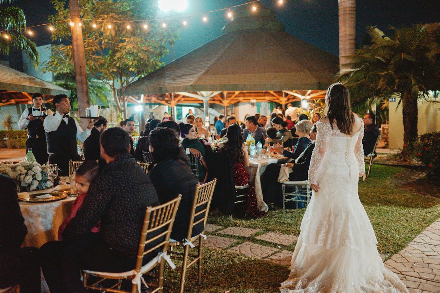 Guests eating at round tables under string lights at an outdoor evening wedding reception, the bride walking past