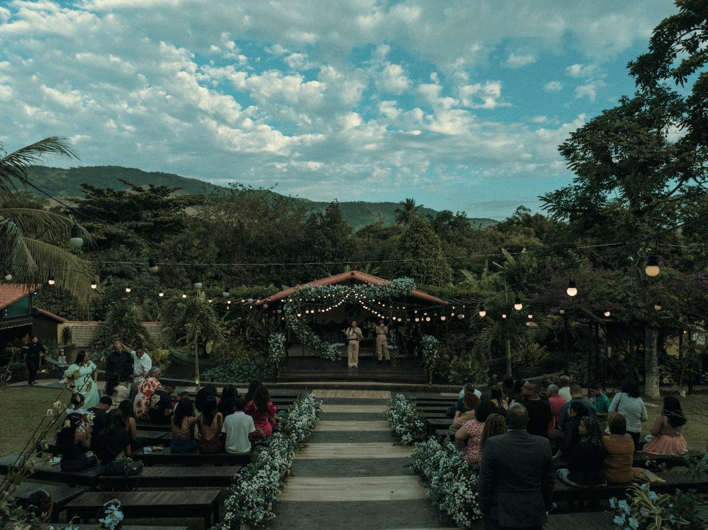 Guests seated on both sides of an aisle lined with white flowers, leading to a stage with greenery and string lights at an outdoor wedding