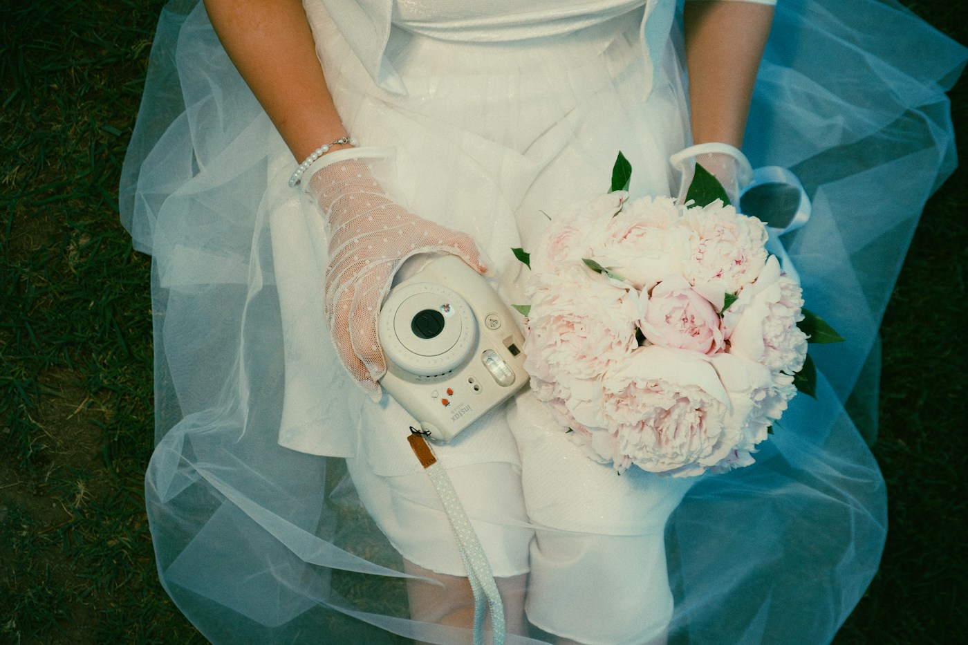 Une mariée en jupe de tulle et gants de dentelle tient un appareil photo instantané blanc et un bouquet de pivoines roses