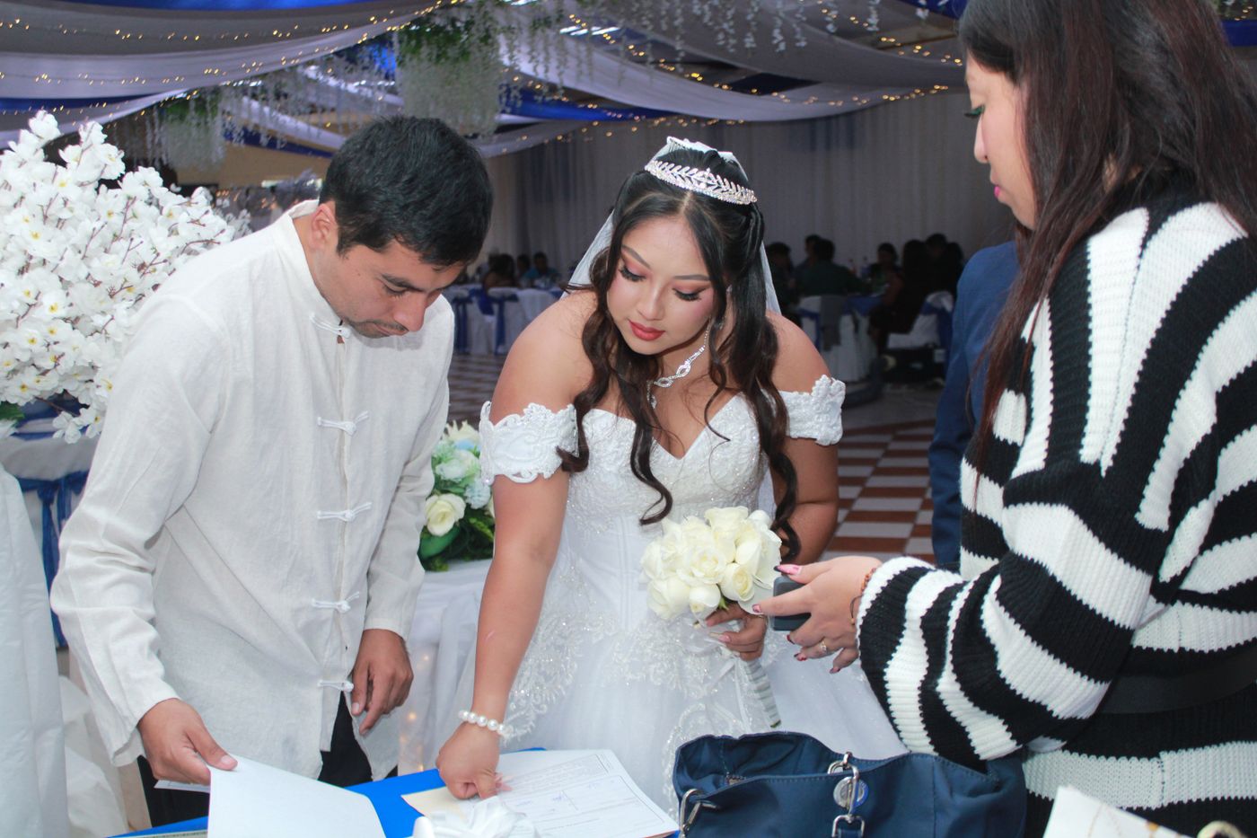 Une invitée en pull rayé tend des papiers à la mariée pendant que le marié lit à une table décorée
