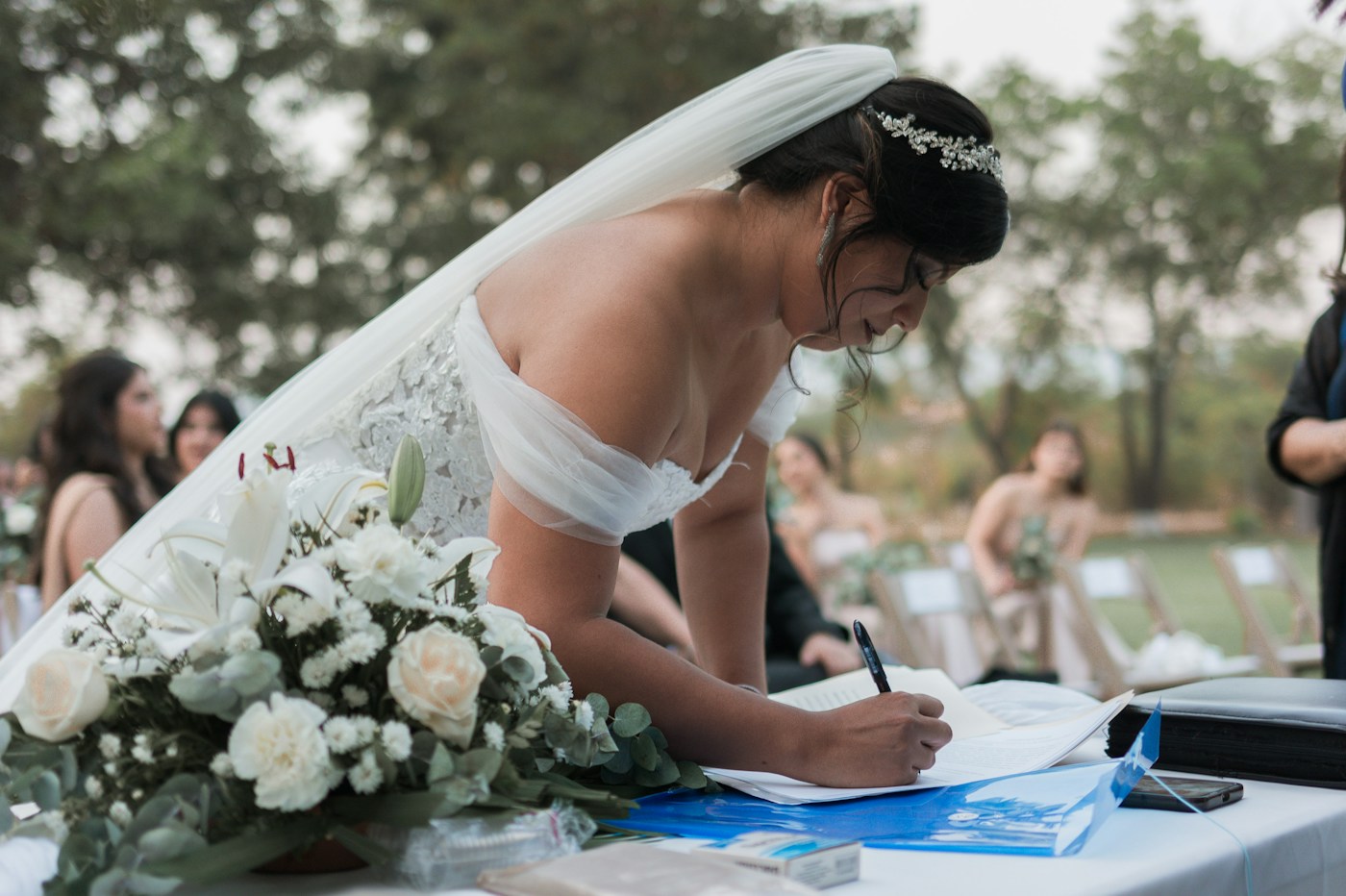 Une mariée en voile se penche sur une table pour écrire pendant que les invités discutent sur la pelouse