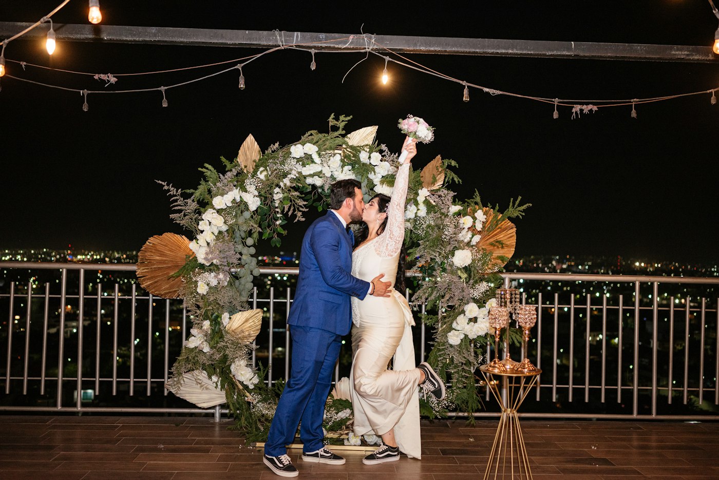 Le marié en costume bleu embrasse la mariée sous une guirlande d'ampoules lors d'un mariage de nuit sur un toit-terrasse