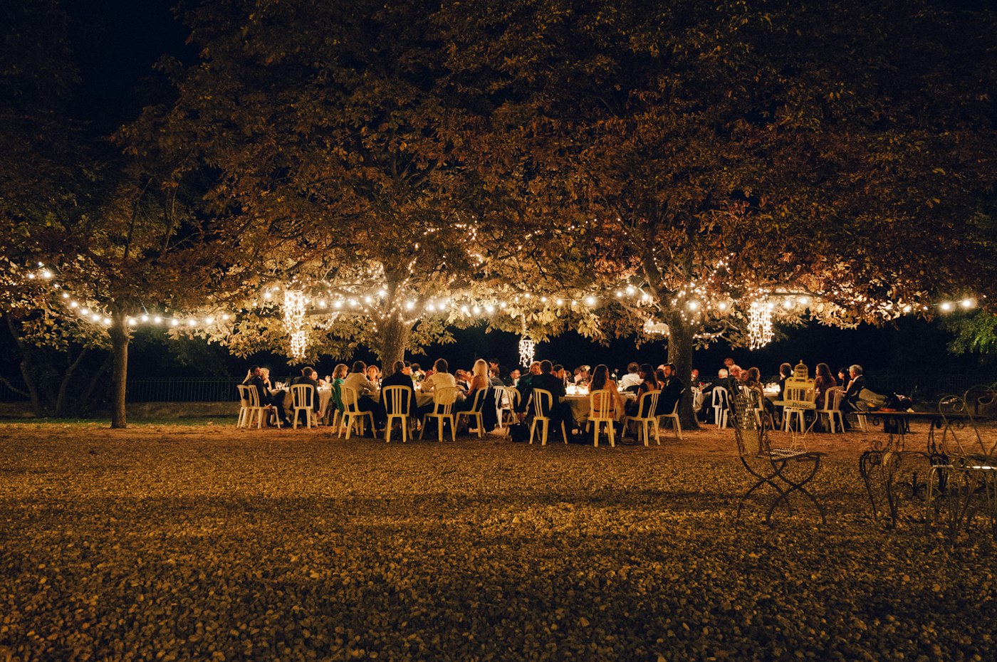 Des invités de mariage dînent la nuit à de longues tables sous de grands arbres garnis de guirlandes d'ampoules chaudes et de grappes de petites lumières
