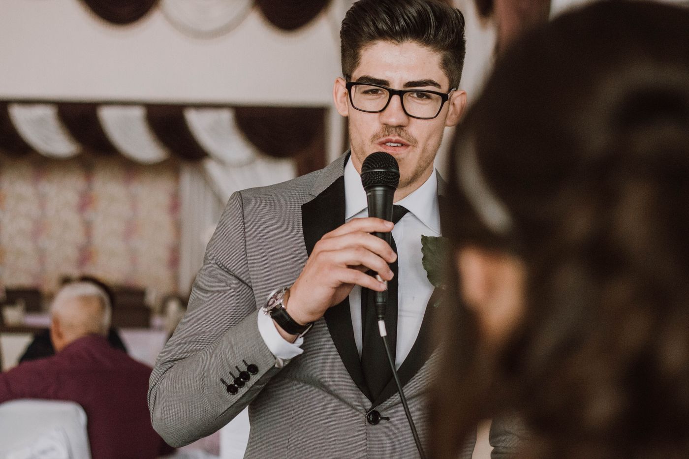 A groomsman in a grey suit with a boutonniere speaks into a handheld microphone at a reception