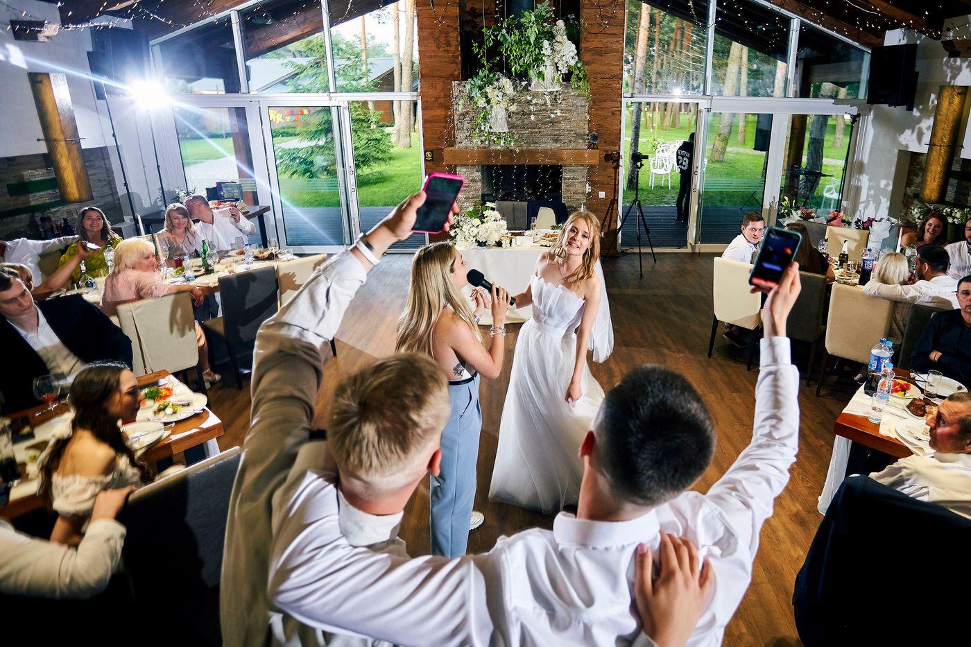 A bridesmaid sings into a microphone to the bride on the dance floor while guests cheer and film on their phones