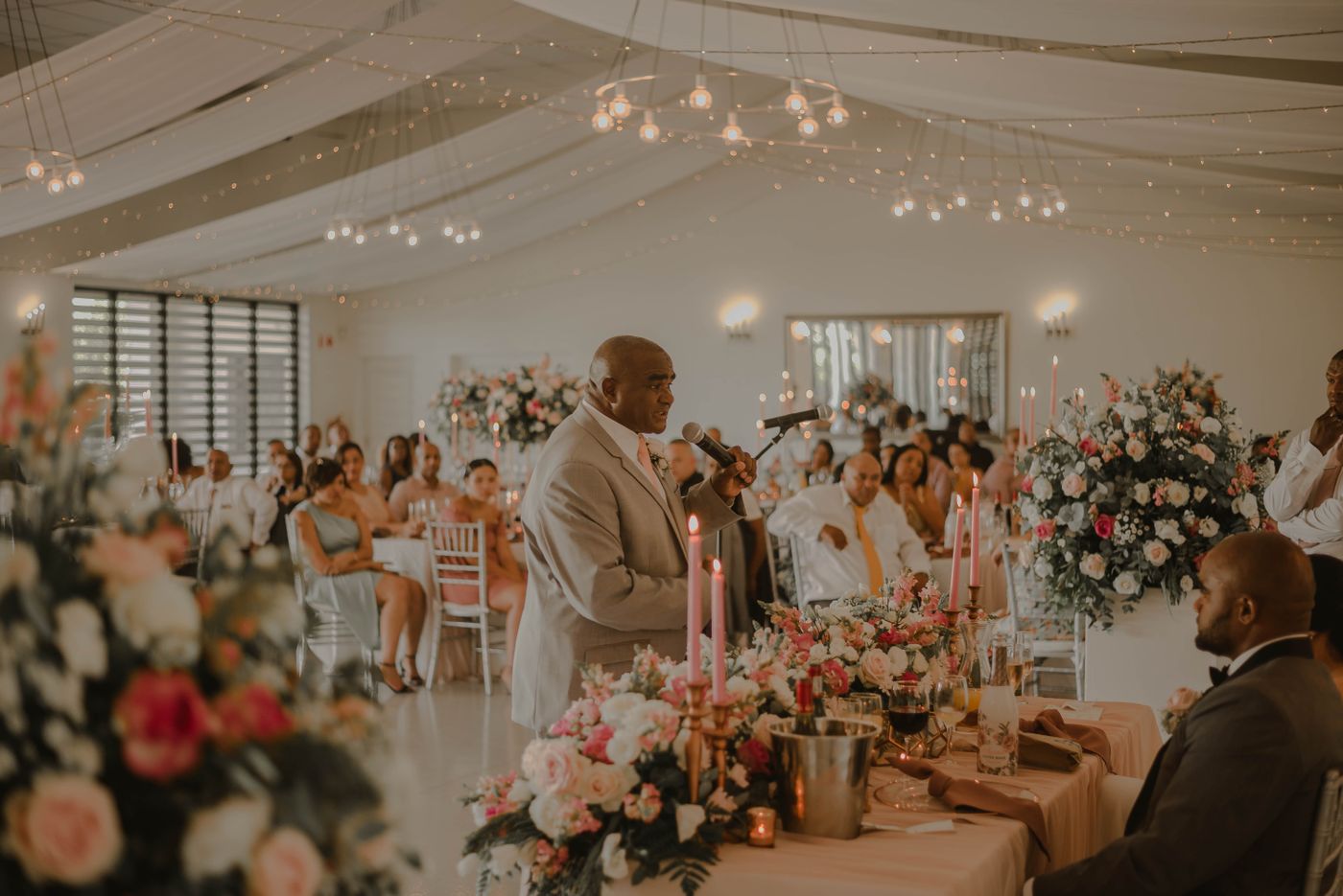 A man in a beige suit speaks into a microphone at a wedding reception with pink flowers, candles and guests listening at their tables