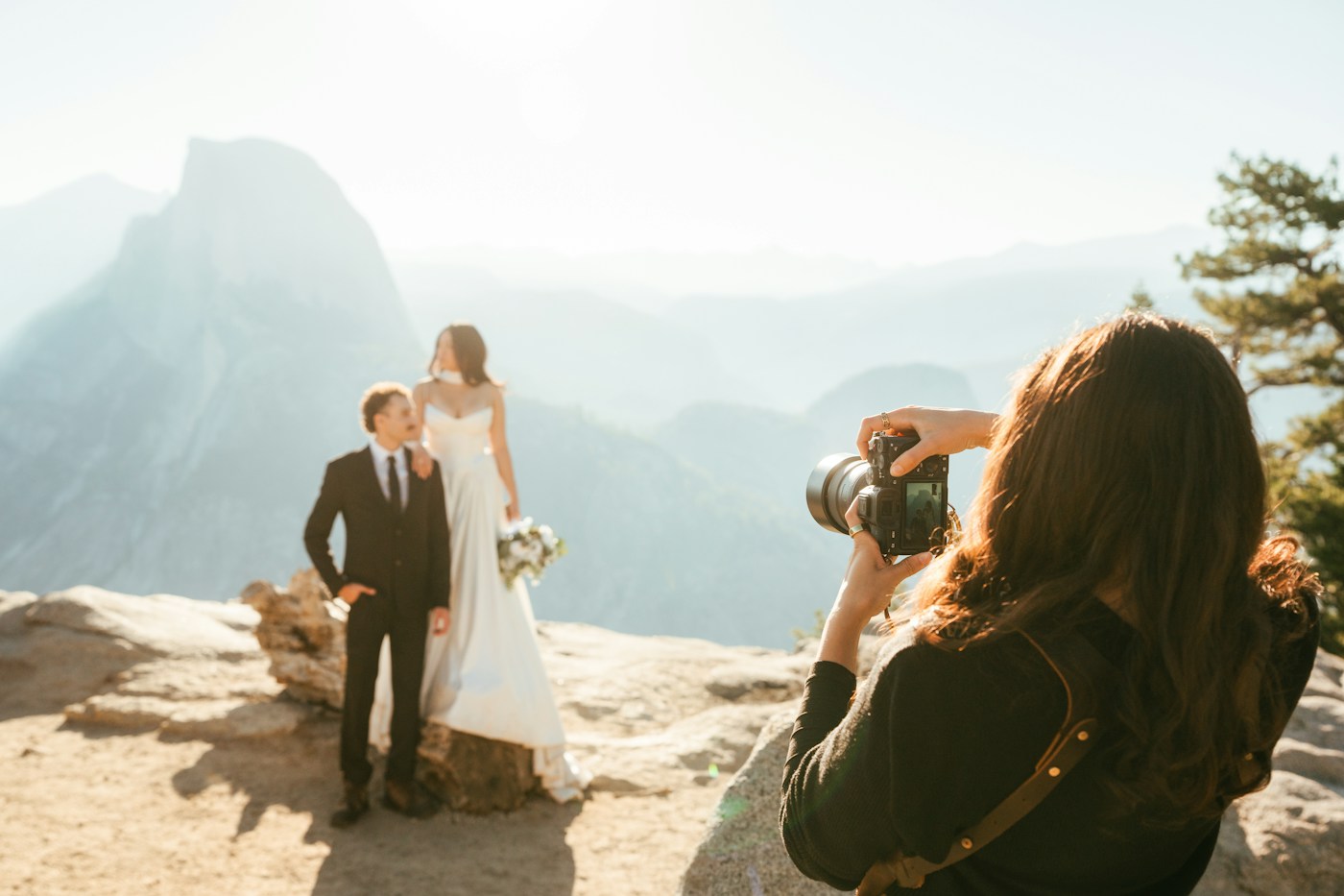 A photographer with a camera photographs a bride and groom on a rocky mountain overlook with granite mountains behind them