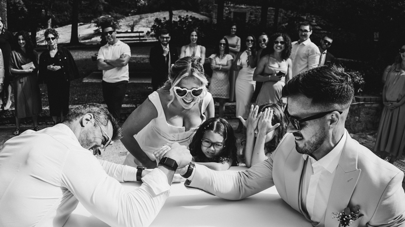 A bride in heart-shaped sunglasses laughs as two men arm wrestle at a table while guests watch in a garden