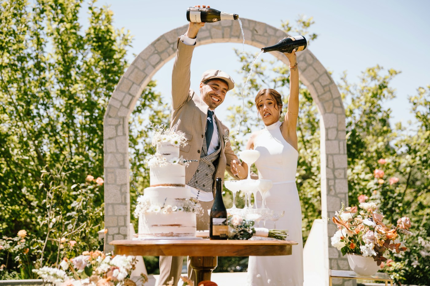 A laughing bride and groom pour champagne into a coupe tower next to a three-tier naked cake under a stone arch in a sunny garden