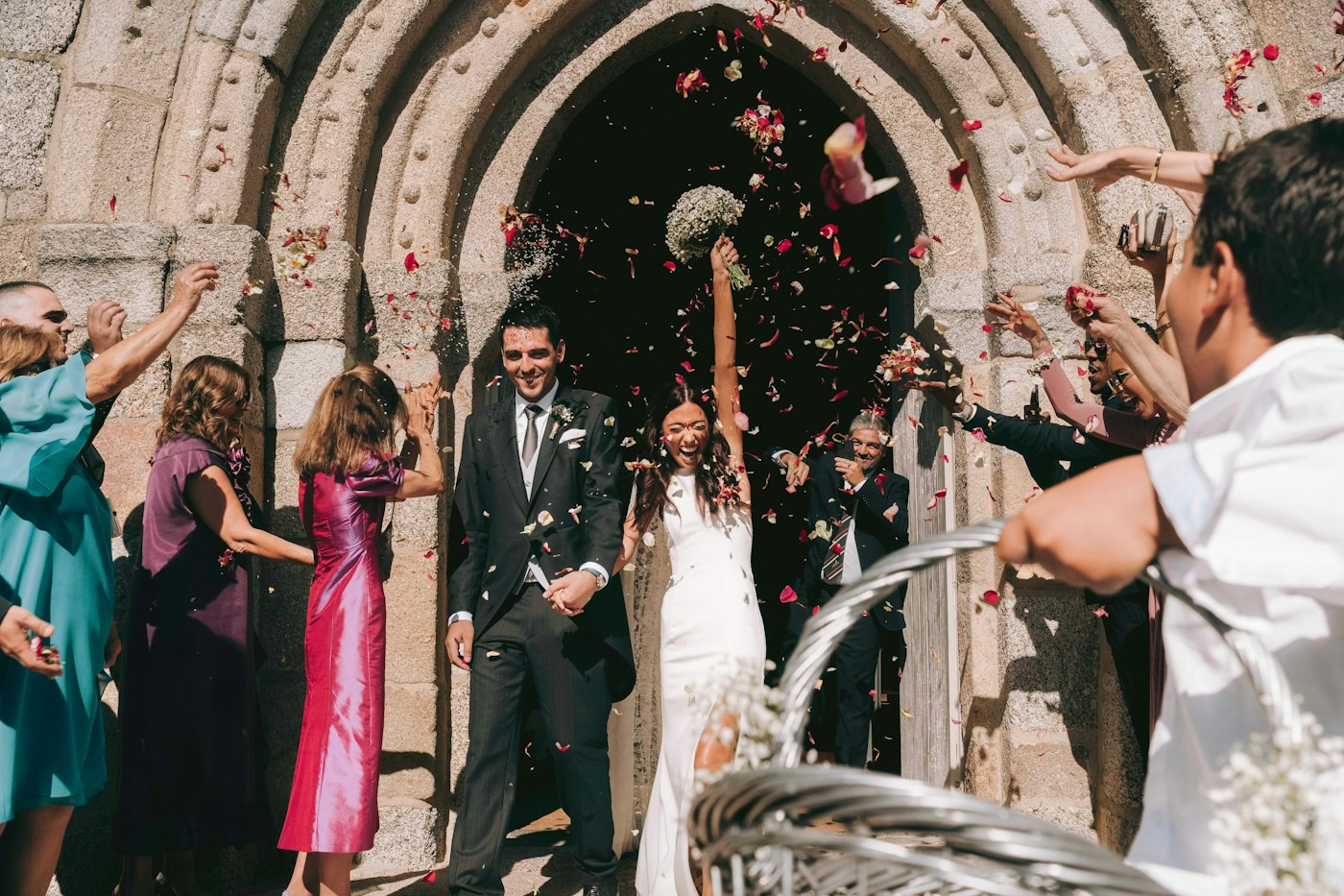 Guests throw red and white petals as the bride and groom walk out of an old stone church, the bride holding a small baby's breath bouquet up high