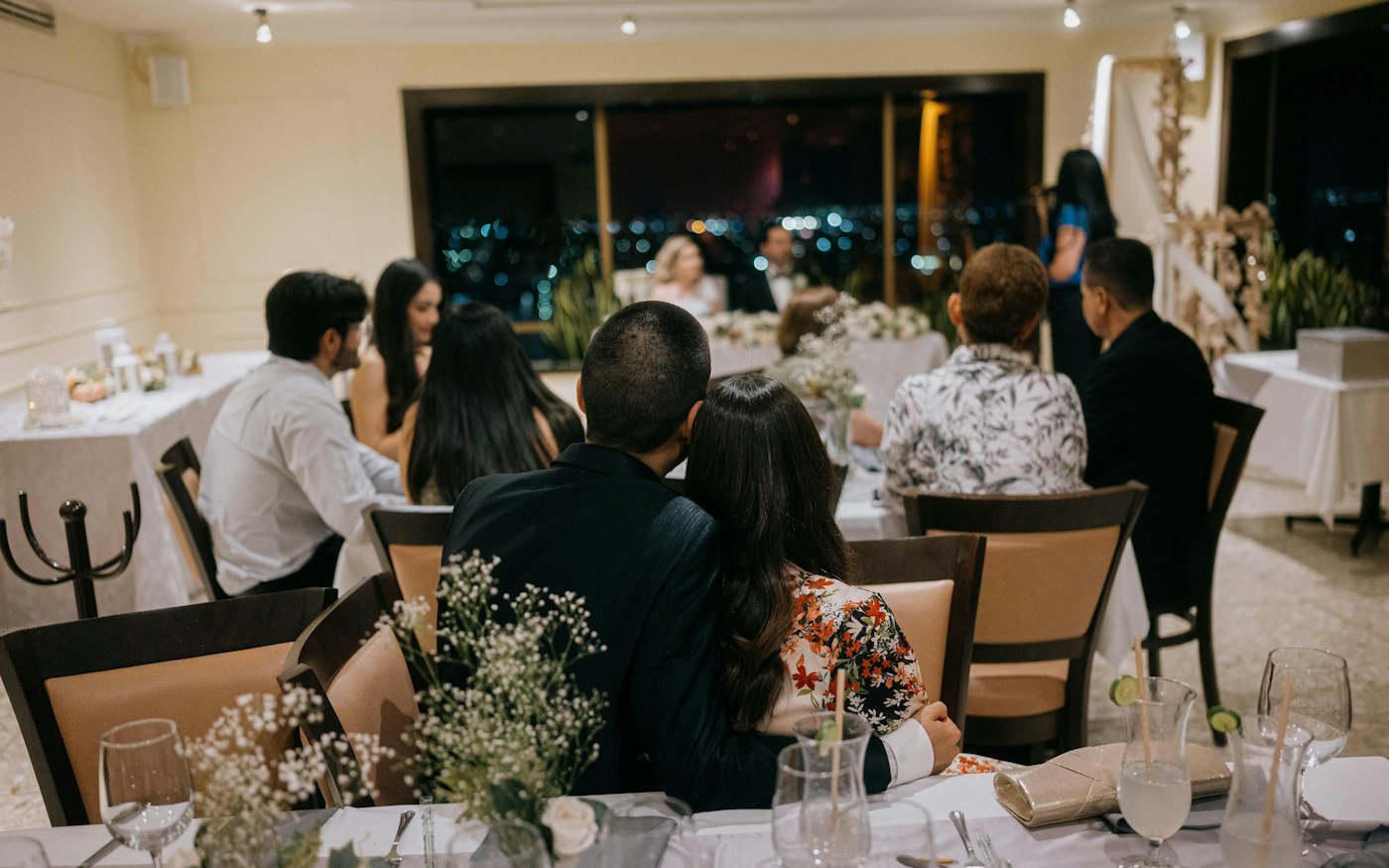 A couple leans into each other at a small reception table with baby's breath in glass vases while guests chat at the next tables