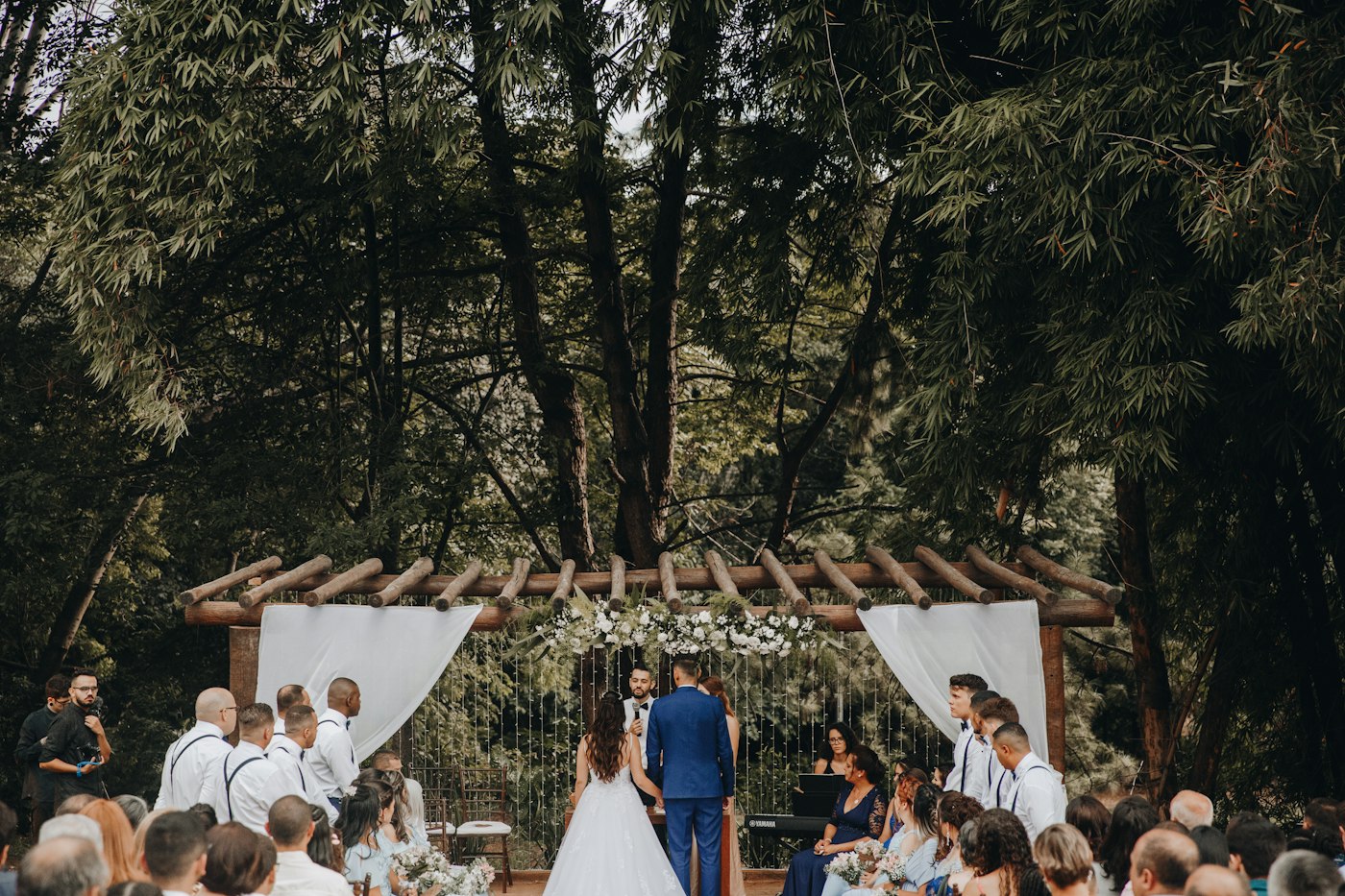 A couple marries under a wooden pergola with white drapes in a wooded garden while guests watch from both sides of the aisle