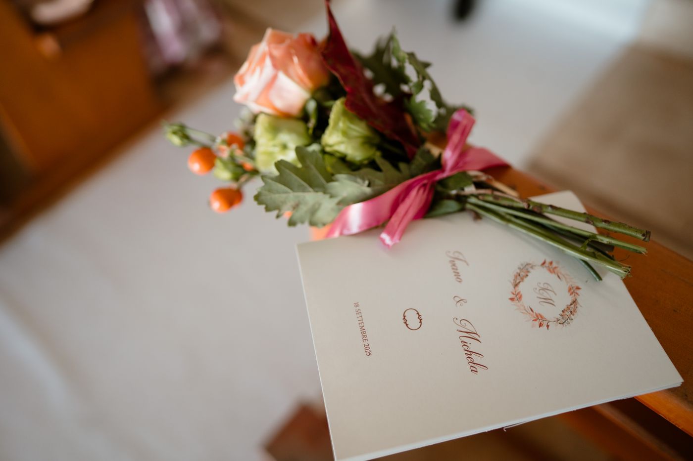 A folded cream wedding program with the couple's names and a monogram wreath rests on a church pew under a small bunch of roses tied with pink ribbon