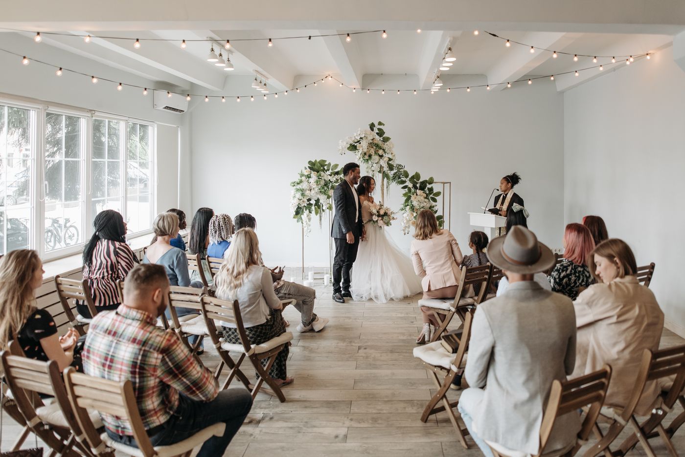Guests on wooden folding chairs watch a couple stand under a white flower arch while the officiant reads at a small lectern