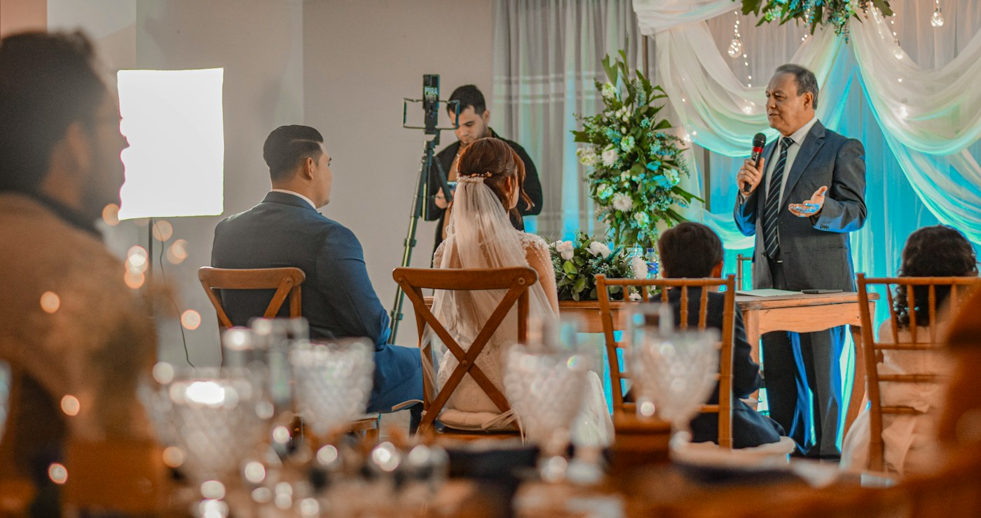 An older guest in a grey suit speaks into a microphone in front of a flower arrangement while the bride and groom listen from their chairs