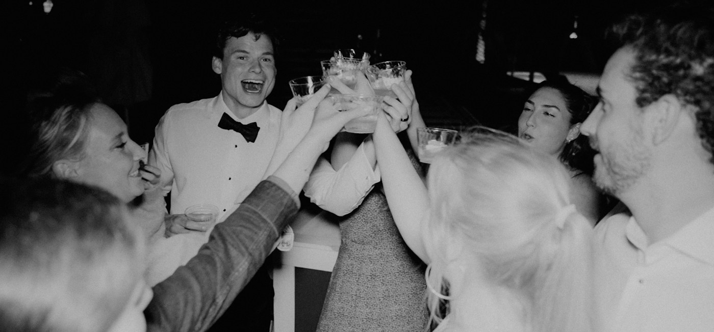 Friends and the groom in a bow tie clink glasses together in a black and white photo at a wedding party