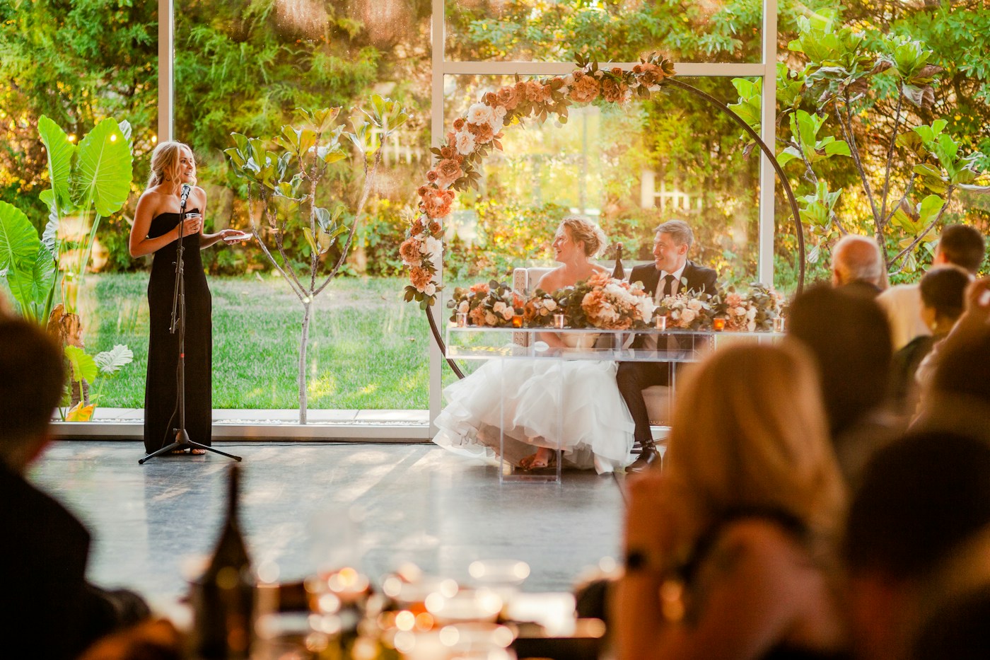A friend in a black dress gives a toast at a microphone while the bride and groom laugh at their flower-arch sweetheart table
