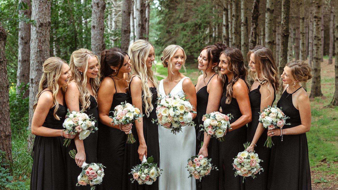 A bride in white laughs with her bridesmaids in black dresses, all holding pink and cream rose bouquets in a pine forest