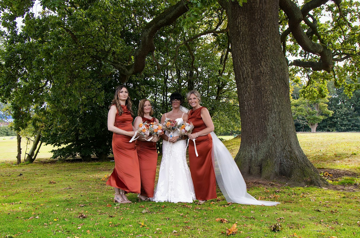 A bride in a lace gown and veil stands with three bridesmaids in rust satin dresses under a large oak tree, all holding orange and cream bouquets