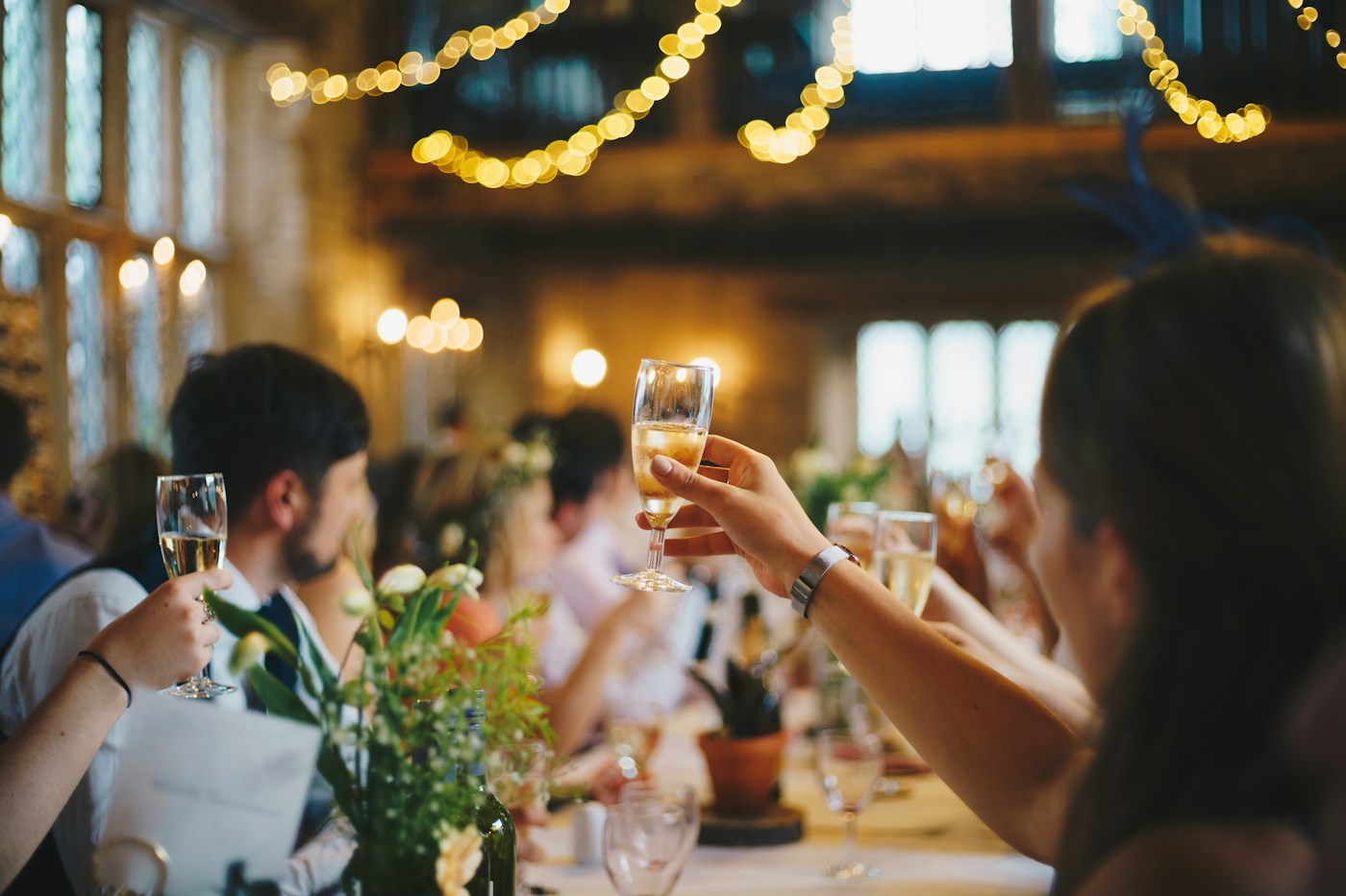 Wedding guests raise glasses of champagne at a long reception table with wildflowers in bottles and fairy lights overhead