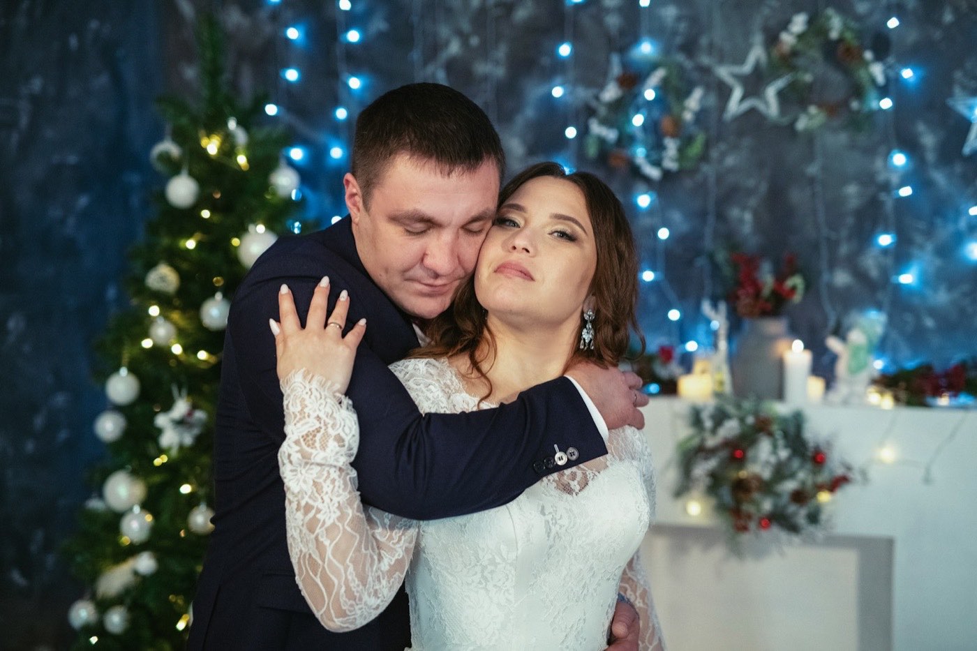 Bride and groom hug in front of a decorated Christmas tree, blue fairy lights and a small candlelit table