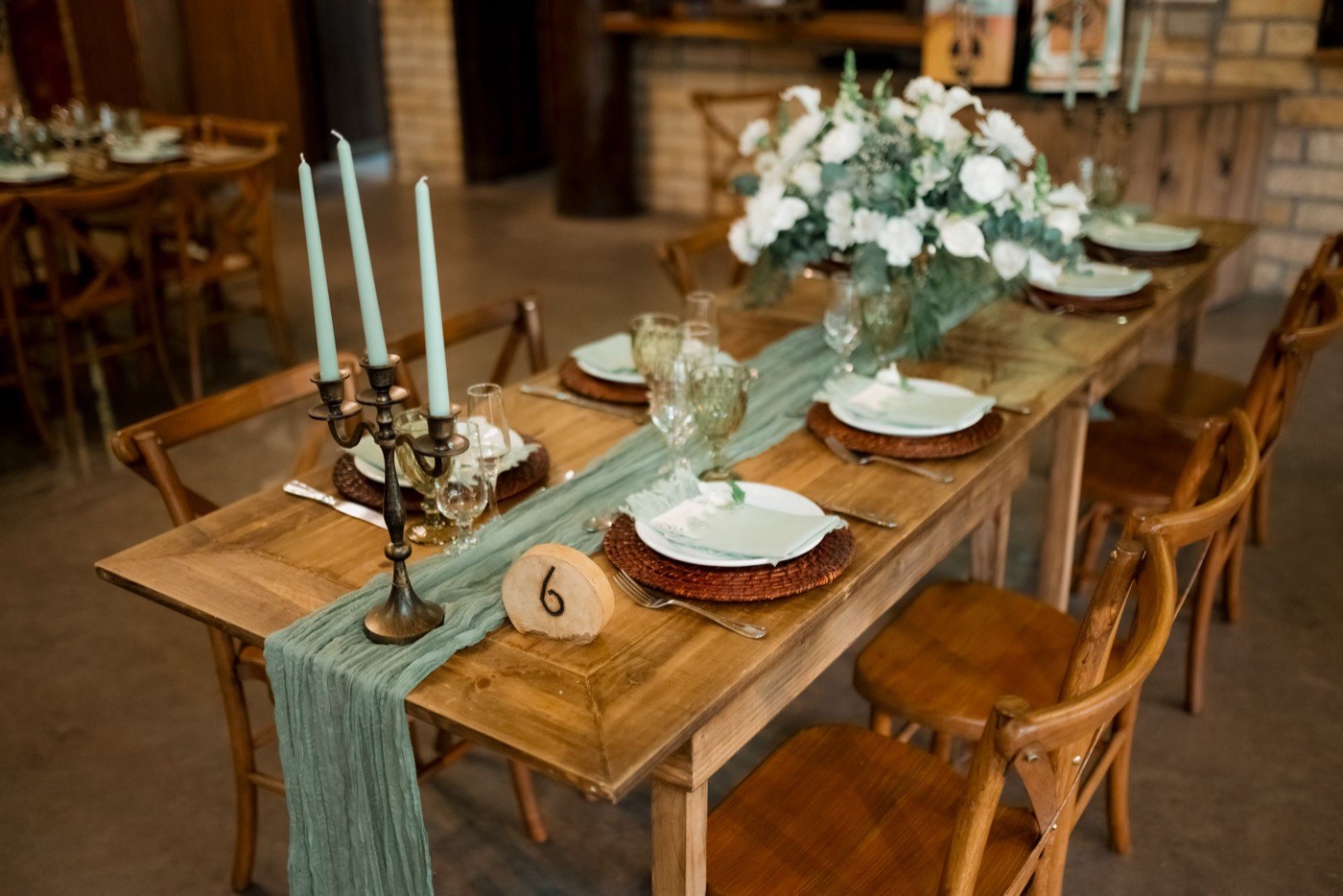 A loosely gathered sage gauze runner down a long wooden wedding table, with candlesticks, white flowers and a wood-slice table number