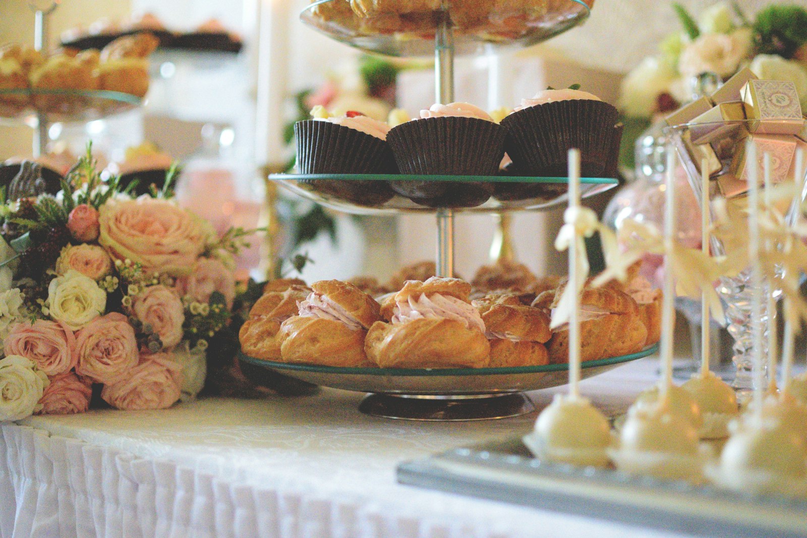 Eine Glas-Etagere mit Cupcakes und Windbeuteln auf einem weiß gedeckten Hochzeits-Dessertbuffet, daneben rosa und cremefarbene Rosen und Cake Pops