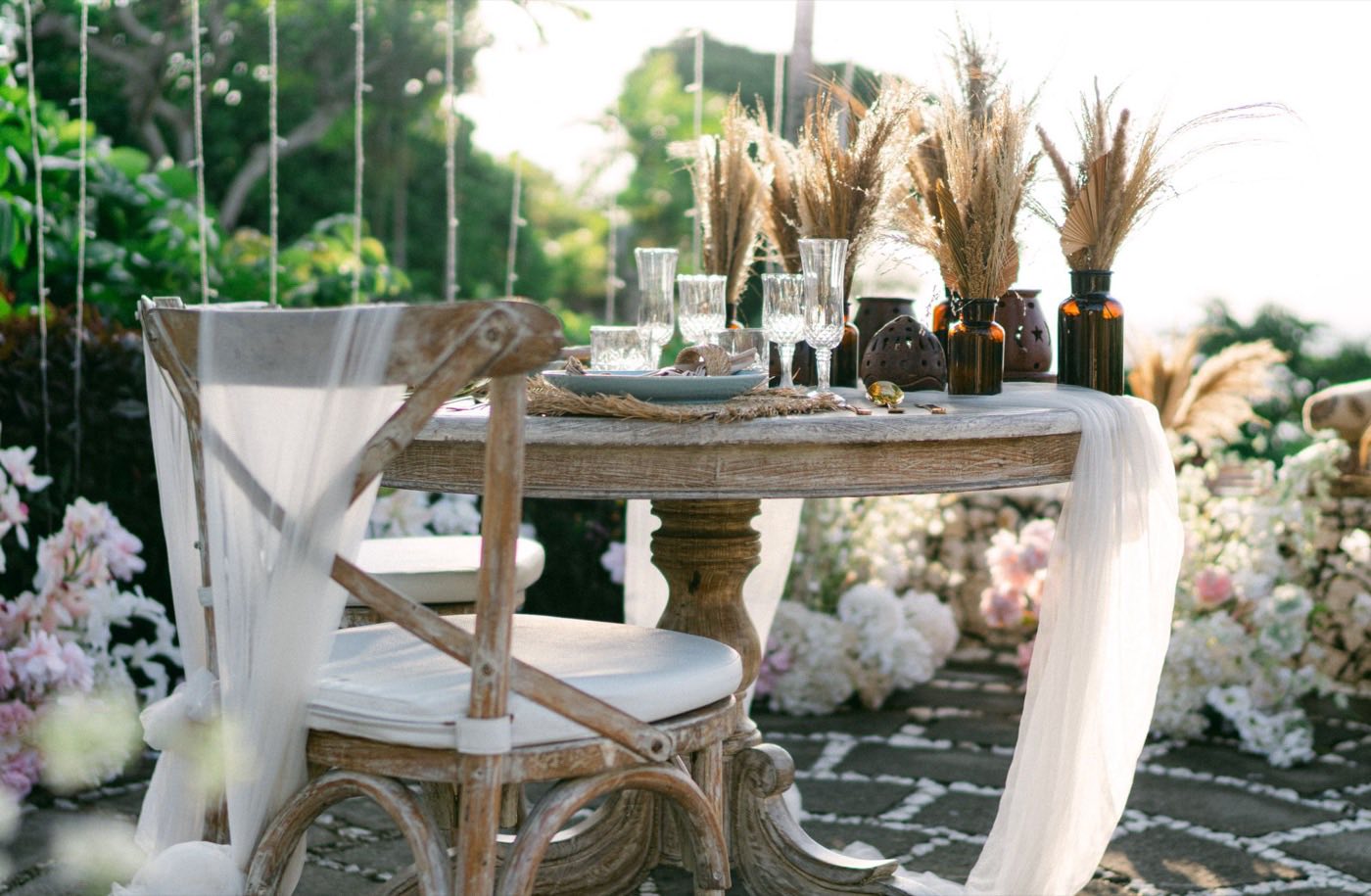Pampas grass plumes in small amber bottles and bud vases on a rustic outdoor wedding table with a white chiffon runner