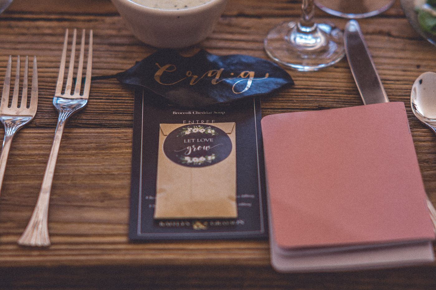 A dark green magnolia leaf with a guest's name written in gold, lying on a menu card at a wooden wedding table