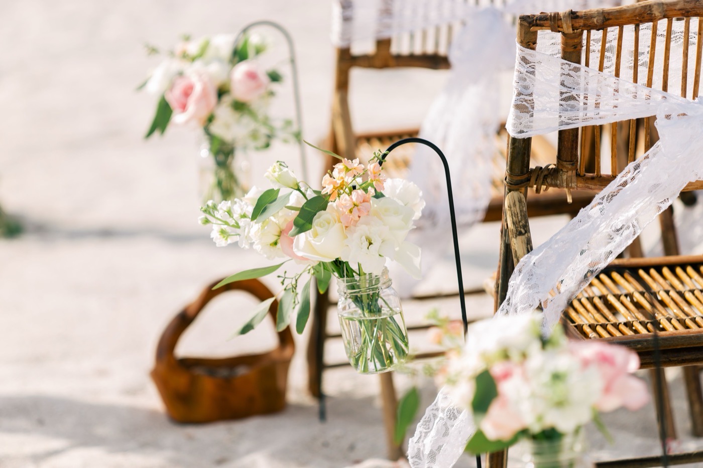 Shepherd hooks lining a beach wedding aisle, each holding a glass jar of pink and white flowers beside the chairs